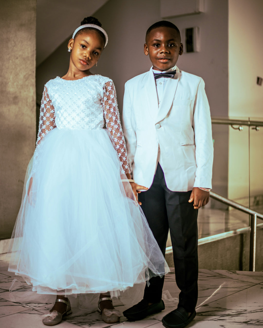 Elegant children's luxury formalwear featuring a white flower girl dress with lace sleeves and a boy's white tuxedo jacket, inspiration by Sewn In The Wood.